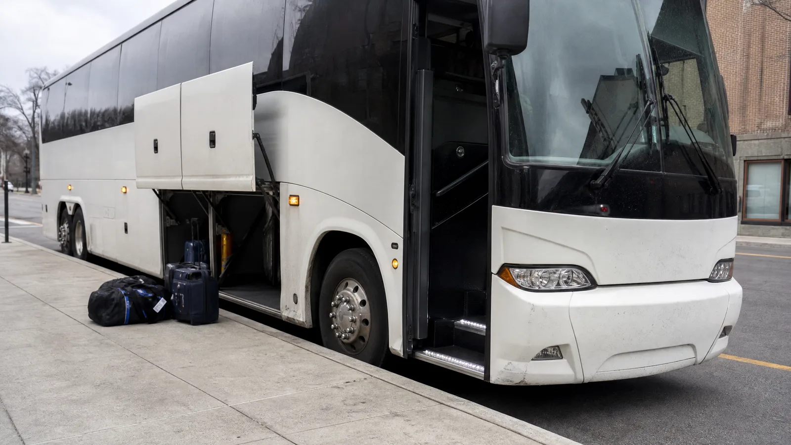 Motor coach parked at a curb with its underfloor luggage bays open and bags waiting on the sidewalk