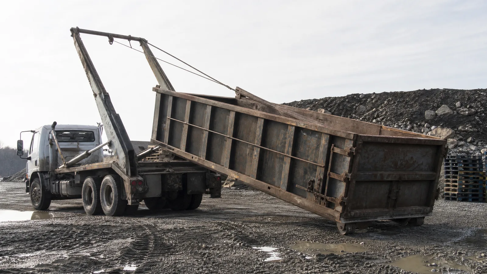 Roll-off truck with its hoist raised lowering a steel container onto gravel at a job site